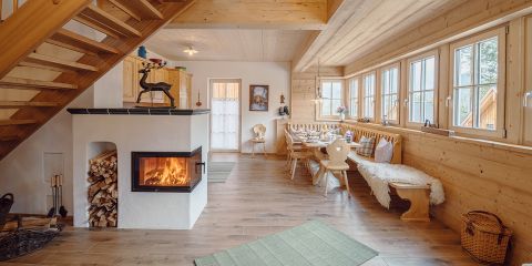 Gemütliches Holzhütten-Interieur mit modernem Kamin, gestapeltem Brennholz, Esstisch mit Stühlen und einer Bank an großen Fenstern. Eine Treppe führt nach oben. Zur Dekoration gehören ein Metallhirsch und gerahmte Kunstwerke.
