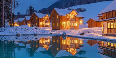 Gemütliche Holzhütten mit schneebedeckten Dächern, warm beleuchtet, spiegeln sich in einem ruhigen Teich. Die Szene spielt vor dem Hintergrund schneebedeckter Hügel und Bäume und schafft in der Dämmerung eine ruhige Winteratmosphäre.
