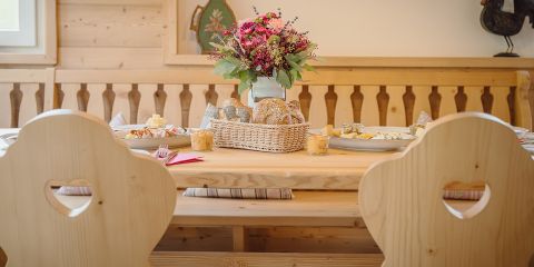 Ein Esstisch aus Holz mit herzförmigen Stühlen in gemütlicher Atmosphäre, dekoriert mit einem Blumengesteck in einer Vase. Ein Korb mit Brot und eine Platte mit Speisen stehen auf dem Tisch, während Sonnenlicht durch ein nahes Fenster hereinfällt.