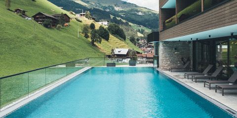 Außenpool mit klarem, blauem Wasser, umgeben von moderner Architektur und Liegestühlen. Mit Blick auf einen malerischen Hügel mit grünem Gras und Holzchalets. Die Berge und Bäume in der Ferne tragen zur ruhigen, malerischen Umgebung bei.