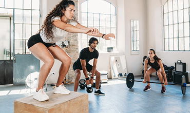 Drei Personen in einem Fitnessstudio führen verschiedene Übungen durch. Eine Person macht Kastensprünge, eine andere Kettlebell-Schwünge und die dritte hebt eine Langhantel. Im Hintergrund sind große Fenster und Fitnessgeräte zu sehen.