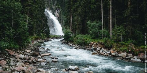 Ein Fluss mit fließendem Wasser führt zu einem Wasserfall, der von dichtem Wald umgeben ist. Die felsigen Flussufer sind von Bäumen gesäumt, und der Wasserfall stürzt eine Klippe hinab und schafft so eine ruhige Naturlandschaft.
