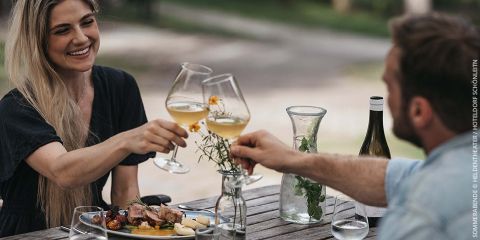 Eine Frau und ein Mann stoßen mit Weingläsern an einem Tisch im Freien an. Vor ihnen stehen Teller mit Essen, daneben eine Karaffe mit Minze und eine Flasche Wein. Beide lächeln, umgeben von einem verschwommenen grünen Hintergrund.