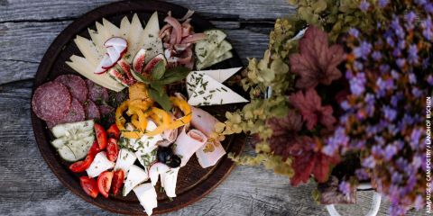 Eine rustikale Holzplatte mit verschiedenen Käsesorten, Salamischeiben, Feigen, Radieschen, gelben Paprikaschoten, Kirschtomaten und Schinken, garniert mit Kräutern. Ein Strauß Hortensien und anderer Blumen verleiht dem Gericht eine farbenfrohe Note.