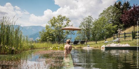 Eine Person steht in einem Naturschwimmteich, umgeben von Grün. In der Nähe befinden sich Liegestühle, ein Sonnenschirm und Bäume. Im Hintergrund sind Berge unter einem teilweise bewölkten Himmel zu sehen.
