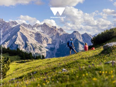 Zwei Wanderer genießen einen malerischen Bergblick unter blauem Himmel mit vereinzelten Wolken. Sie stehen auf einem grasbewachsenen Hügel mit Wildblumen und blicken auf felsige Gipfel im Hintergrund. Das Logo „Wanderhotels“ ist oben im Bild zu sehen.