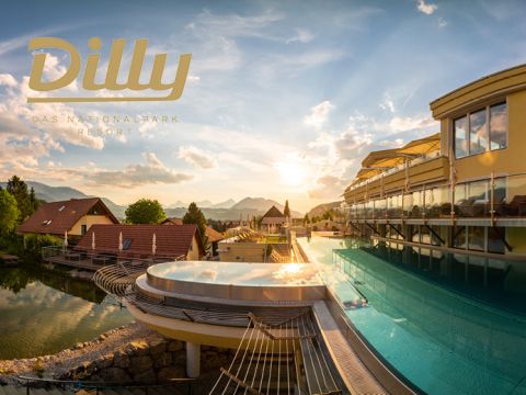 Außenpool eines Resorts mit Glasgeländer vor der malerischen Kulisse von Bergen und einem farbenprächtigen Sonnenuntergang. Die Gebäude des Resorts verfügen über große Fenster, im Vordergrund sind üppiges Grün und ein kleiner Teich zu sehen.