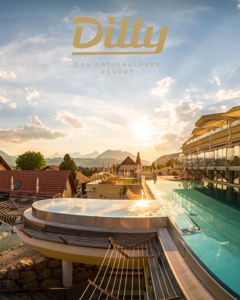 Infinity-Pool in einem Resort mit Blick auf eine malerische Berglandschaft bei Sonnenuntergang. Moderne Architektur mit Glas- und Holzelementen, weitere Strukturen sichtbar. Das Logo „Dilly Das Nationalpark Resort“ steht oben. Der Himmel ist von sanften Wolken geprägt und strahlt warmes Licht aus.
