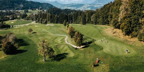 Luftaufnahme eines weitläufigen Golfplatzes mit üppig grünen Fairways und Sandbunkern. Mehrere Golfer spielen auf dem Platz, umgeben von Bäumen und fernen Hügeln. Der Himmel ist klar und bietet eine malerische Landschaft.
