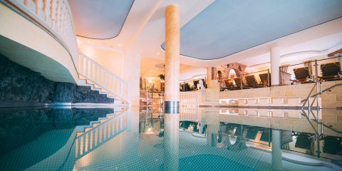 Luxuriöser Innenpoolbereich mit einer Säule in der Mitte, umgeben von eleganter Architektur. Liegestühle säumen den Bereich, und sanfte Beleuchtung schafft eine entspannende Atmosphäre. Die Decke und die Spiegelungen verleihen der ruhigen Szene Tiefe.
