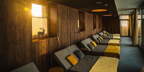 Ein gemütlicher Ruheraum ist mit mehreren in einer Reihe angeordneten Loungesesseln ausgestattet, die jeweils mit einem grauen Kissen und einer gelben Decke ausgestattet sind. Warmes Sonnenlicht fällt durch ein Fenster mit Holzjalousien und beleuchtet die Holzwände sowie eine dekorative Rentiersilhouette.