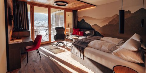 Ein modernes Hotelzimmer mit einem gemütlichen Bett, einem Schreibtisch mit rotem Stuhl und einem schwarzen Loungesessel neben einer Glastür, die zum Balkon führt. Das Zimmer verfügt über ein Wandgemälde mit Bergmotiven und große Fenster mit malerischer Aussicht.