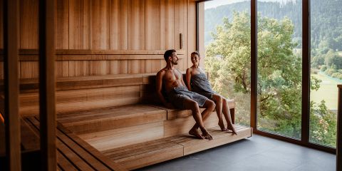 Zwei Personen entspannen auf Holzbänken in einer geräumigen, hellen Sauna. Einer trägt ein Handtuch, der andere ist teilweise zugedeckt. Große Fenster geben den Blick auf Bäume und Hügel im Hintergrund frei.