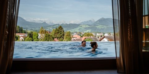 Zwei Personen schwimmen in einem Infinity-Pool mit Blick auf die Berge und ein malerisches Dorf im Hintergrund. Offene Vorhänge rahmen die Szenerie ein und suggerieren einen Blick aus einem Fenster oder von einer Terrasse.