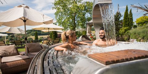 Ein Mann und eine Frau entspannen in einem Whirlpool im Freien mit Wasserfall, umgeben von Bäumen und Liegestühlen mit Sonnenschirmen. Die Umgebung ist sonnig und ruhig und bietet einen Blick auf eine grüne Landschaft.