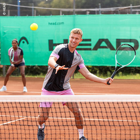 Ein Mann in Sportkleidung bereitet sich darauf vor, mit seinem Schläger einen Tennisball auf einem Sandplatz zu schlagen. Eine Frau steht im Hintergrund und beobachtet aufmerksam. Hinter ihnen ist ein grünes Banner mit dem Schriftzug „HEAD“ zu sehen.