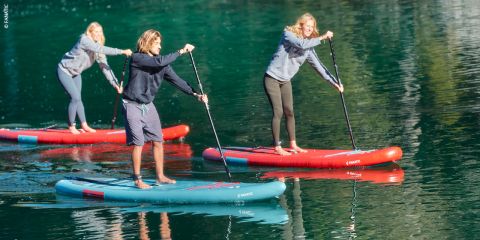 Drei Personen paddeln auf einem ruhigen, klaren Gewässer. Sie balancieren auf roten und blauen Paddleboards und tragen legere Kleidung. Das grüne Wasser spiegelt ihre Bilder wider. Die Szene ist hell und sonnig und lässt auf einen schönen Tag schließen.