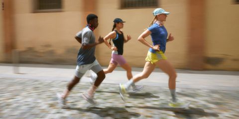 Drei Menschen joggen auf einer Kopfsteinpflasterstraße. Ein Mann in grauem Hemd und zwei Frauen in Sportkleidung, eine mit blauem Hemd und gelben Shorts, die andere in Schwarz und Pink. Sie tragen Mützen und Stirnbänder, sind konzentriert und in Bewegung.