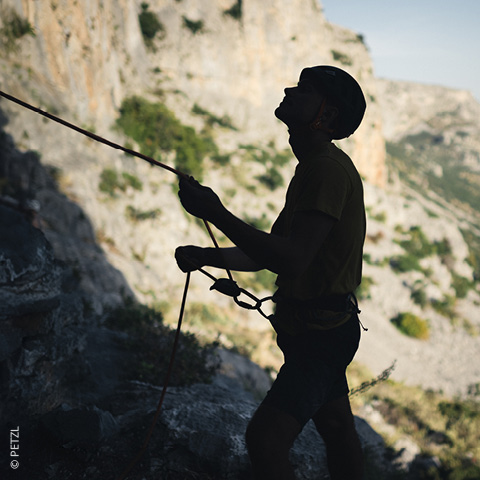 Silhouette einer Person mit Helm und Kletterseil vor einer felsigen Landschaft. Im Hintergrund sind sonnenbeschienene Klippen und grüne Flecken zu sehen.