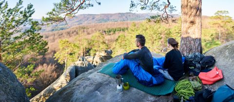 Ein Paar sitzt in Schlafsäcke gehüllt auf einem großen Felsen und blickt auf eine malerische Wald- und Berglandschaft. In der Nähe steht ein kleiner Campingplatz mit Tassen und Wasserkocher. Bäume und ein weiter Himmel runden den ruhigen Anblick ab.