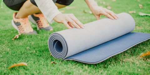 Eine Person kniet im Gras und rollt eine zweifarbige Yogamatte mit grauen und blauen Seiten zusammen. Sie trägt einen cremefarbenen Pullover, eine schwarze Hose und orangefarbene Turnschuhe. Trockene Blätter liegen verstreut auf dem Gras.