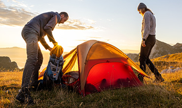Ein Mann und eine Frau bauen bei Sonnenuntergang ein leuchtend orangefarbenes Zelt auf einem grasbewachsenen Hügel auf. Der Mann packt seinen Rucksack, während die Frau zusieht. Der Himmel ist mit zarten Wolken bedeckt und hat einen goldenen Farbton.