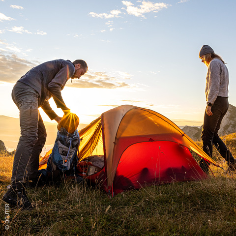 Zwei Personen bauen bei Sonnenuntergang ein rotes Zelt auf einem grasbewachsenen Hügel auf. Eine Person packt einen Rucksack, die andere steht daneben. Der Himmel ist teilweise bewölkt und taucht die Szene in einen warmen Schein. Im Hintergrund sind Berge zu sehen.
