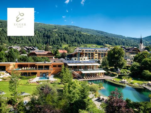 Malerische Aussicht auf ein Luxusresort, umgeben von üppigem Grün und Bergen. Es gibt einen kleinen Teich, Sitzgelegenheiten im Freien und mehrere Gebäude, die modern und rustikal zugleich wirken. Im Hintergrund ist ein Kirchturm zu sehen. Darüber strahlend blauer Himmel.