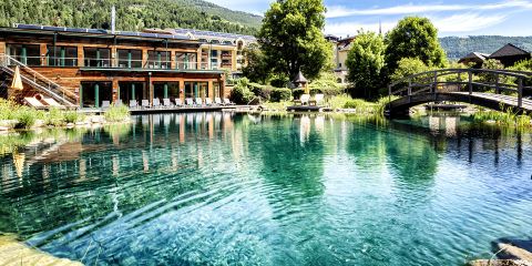 Ein malerischer Blick auf ein Gebäude mit großen Fenstern an einem natürlichen Swimmingpool, umgeben von Grün und Bäumen. Eine Holzbrücke führt über einen Teil des Pools, und auf der Terrasse stehen Liegestühle unter einem klaren blauen Himmel.