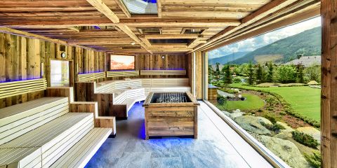 Eine geräumige Holzsauna mit Sitzstufen und stimmungsvoller Beleuchtung verfügt über ein großes Fenster mit Blick auf einen üppigen Garten und die Berglandschaft. Ein zentraler, holzverkleideter Steinofen trägt zur ruhigen und natürlichen Atmosphäre bei.