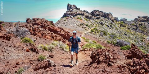 Eine Person in Wanderkleidung geht einen felsigen Pfad mit rötlichem Vulkangestein und spärlichem Grün entlang. Dahinter sind eine raue Landschaft und ein klarer blauer Himmel zu sehen. Sie trägt eine Mütze, eine Sonnenbrille und einen Rucksack.