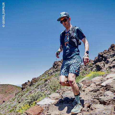 Ein Mann mit Mütze und Sonnenbrille läuft unter strahlend blauem Himmel auf einem steinigen Pfad. Er trägt ein blaues Hemd und karierte Shorts, einen Rucksack und eine Armbanduhr und ist von einer rauen Landschaft mit spärlicher Vegetation umgeben.