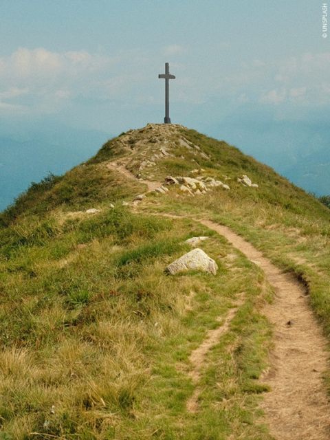 Eine grasbewachsene Hügelkuppe mit einem Feldweg, der zu einem großen Kreuz auf dem Gipfel führt. Im Hintergrund ist der Himmel teilweise bewölkt.