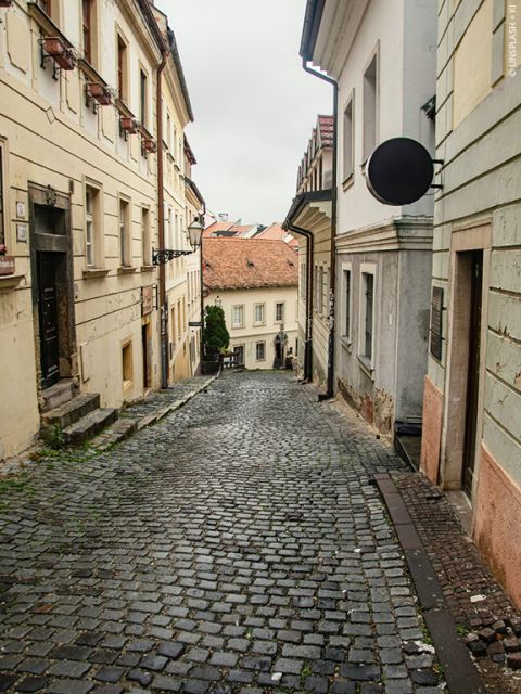Schmale Kopfsteinpflasterstraße in einer europäischen Stadt, gesäumt von alten Gebäuden mit verblassten Mauern und Fensterläden. Die Straße führt bergab und schafft unter bewölktem Himmel eine malerische, historische Atmosphäre.