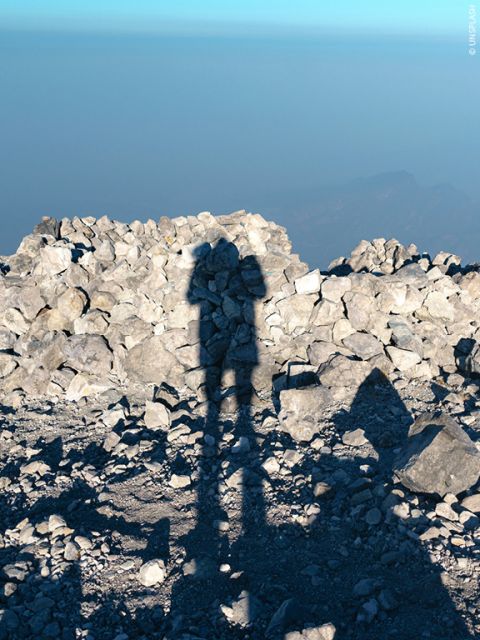 Schatten einer Person mit einer Kamera auf felsigem Gelände, vor dem Hintergrund verstreuter Steine und eines dunstigen, fernen Horizonts unter einem klaren blauen Himmel.