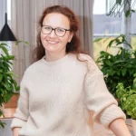Eine lächelnde Person mit langen Haaren und Brille steht in einem leichten Pullover im Innenbereich. Sie ist von grünen Pflanzen umgeben, im Hintergrund fällt natürliches Licht durch ein Fenster.