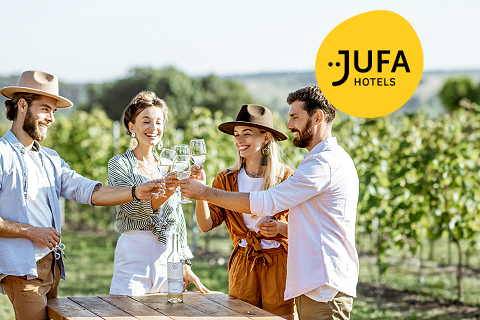 Vier Personen stehen draußen in einem Weinberg und stoßen mit Weingläsern an. Sie lächeln und sind leger gekleidet, tragen Hüte. Vor ihnen steht ein Picknicktisch mit einer Weinflasche. Oben rechts ist das Logo der JUFA Hotels zu sehen.