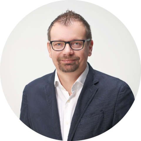 Ein Mann mit kurzen Haaren und Brille trägt ein dunkles Jackett über einem weißen Hemd mit Kragen und steht vor einem schlichten hellen Hintergrund.