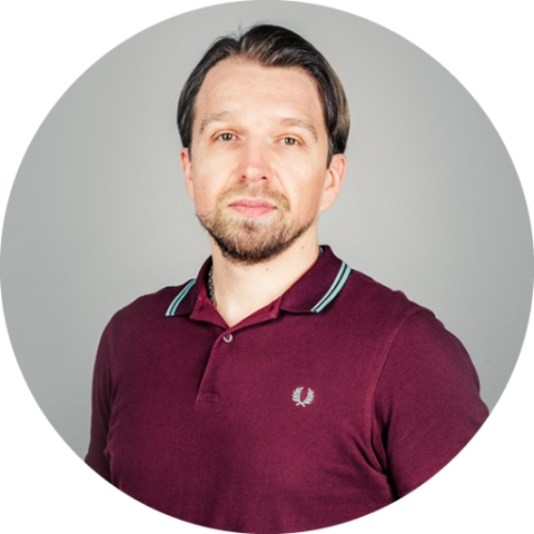 Porträt einer Person mit kurzen dunklen Haaren und Bart. Sie trägt ein burgunderfarbenes Poloshirt und steht vor einem schlichten grauen Hintergrund. Ihr Gesichtsausdruck ist neutral und sie blickt nach vorne.