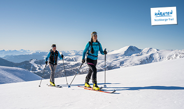 Zwei Skifahrer mit Stöcken und blauen Jacken fahren an einem sonnigen Tag eine verschneite Loipe entlang, im Hintergrund sind Berggipfel zu sehen. Oben rechts steht ein Schild mit der Aufschrift „Kärnten Nockberge-Trail“.