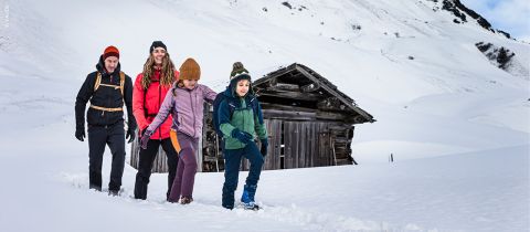 Eine Gruppe von vier Personen in farbenfroher Winterkleidung geht durch den Schnee. Im Hintergrund ist eine Holzhütte zu sehen. Unter einem klaren blauen Himmel sind schneebedeckte Berge zu sehen.