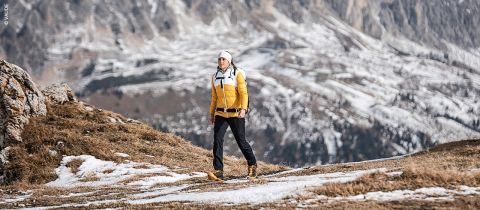 Eine Person in gelber Jacke und weißem Hut wandert auf einem verschneiten Bergpfad. Die felsige Landschaft im Hintergrund ist teilweise mit Schnee bedeckt.