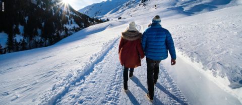 Ein Paar geht Hand in Hand auf einem verschneiten Bergpfad. Sie tragen Winterjacken und Mützen. Die Sonne scheint hell über die schneebedeckte Landschaft, links sind Kiefern zu sehen, im Hintergrund Berge.