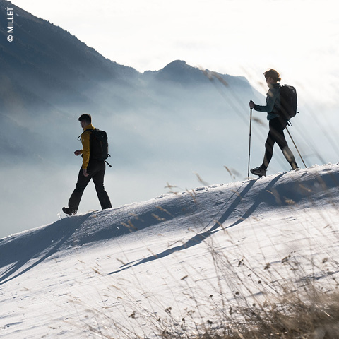 Zwei Wanderer mit Rucksäcken und Stöcken wandern über einen schneebedeckten Hang. Die Sonne wirft lange Schatten und hebt das strukturierte Gelände hervor. Im Hintergrund ist eine neblige Bergkette zu sehen.