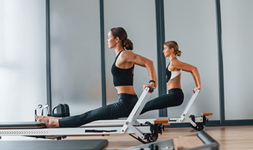 Zwei Frauen in Sportkleidung machen in einem hellen Fitnessstudio Übungen an Pilates-Reformern. Sie strecken ihre Beine und führen eine Trizeps-Dip-ähnliche Bewegung aus, um Kraft und Gleichgewicht zu demonstrieren. Im Hintergrund steht eine Tasche.
