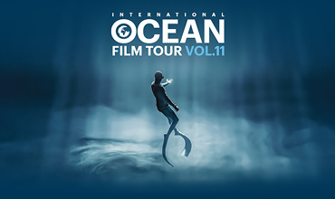 Ein Taucher mit Flossen und Neoprenanzug schwimmt unter Wasser in einer surrealen, blau beleuchteten Meeresszene. Oben steht „International Ocean Film Tour Vol. 11“. Lichtstrahlen durchdringen das Wasser und schaffen eine traumhafte Atmosphäre.
