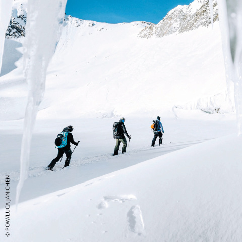 Drei Personen wandern durch eine verschneite Landschaft, umgeben von eisigen Klippen. Sie sind mit Winterausrüstung ausgestattet, darunter Rucksäcke und Trekkingstöcke. Der Himmel ist klar und blau und bildet einen Kontrast zum weißen Schnee.