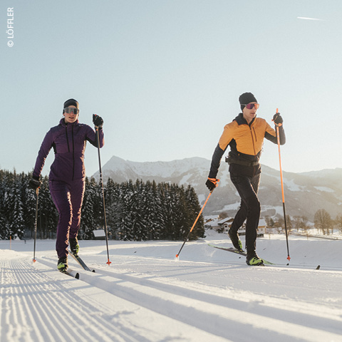 Zwei Personen laufen auf einer präparierten Loipe mit schneebedeckten Bergen und immergrünen Bäumen im Hintergrund. Sie tragen Wintersportausrüstung, darunter Mütze und Handschuhe, unter einem klaren blauen Himmel.