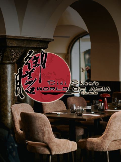 Ein gemütliches asiatisches Restaurant mit rundem Esstisch und gepolsterten Stühlen. An der Wand prangt ein roter Kreis mit schwarzen asiatischen Schriftzeichen und dem Text „World of Asia, à la carte“. Daneben steht eine dekorative Säule.