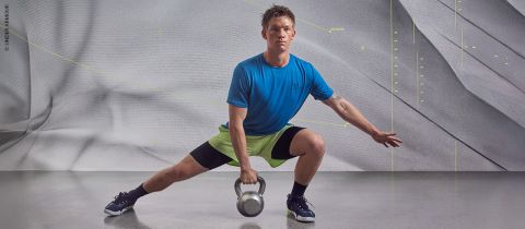 Eine Person in blauem Shirt und grünen Shorts macht einen seitlichen Ausfallschritt und hält dabei eine Kettlebell. Im Hintergrund ist eine strukturierte graue Wand mit dezenten Mustern und Linien zu sehen.